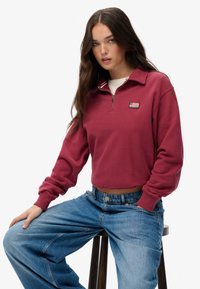 Bordeaux kwart-rits sweatshirt met een kraag, voorzien van een klein logo-embleem. In combinatie met lichtblauwe wijduitlopende jeans van gestructureerde stof.