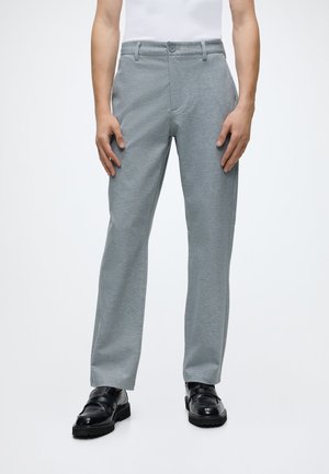 Homme portant un pantalon droit gris clair, des chaussures noires et une chemise blanche, debout devant un fond clair uni.