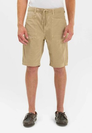 Homme portant un short beige mi-genou avec des poches plaquées à l'avant et des chaussures marron décontractées, debout devant un fond blanc uni.