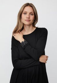 Kobieta z d�ługimi blond włosami, ubrana w czarną bluzkę z długim rękawem i masywną czarną bransoletkę, pozująca na jednolitym jasnym tle.