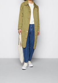 Cappotto lungo imbottito verde oliva con colletto, bottoni a scatto dorati e una delicata texture, abbinato a jeans blu e sneakers bianche.