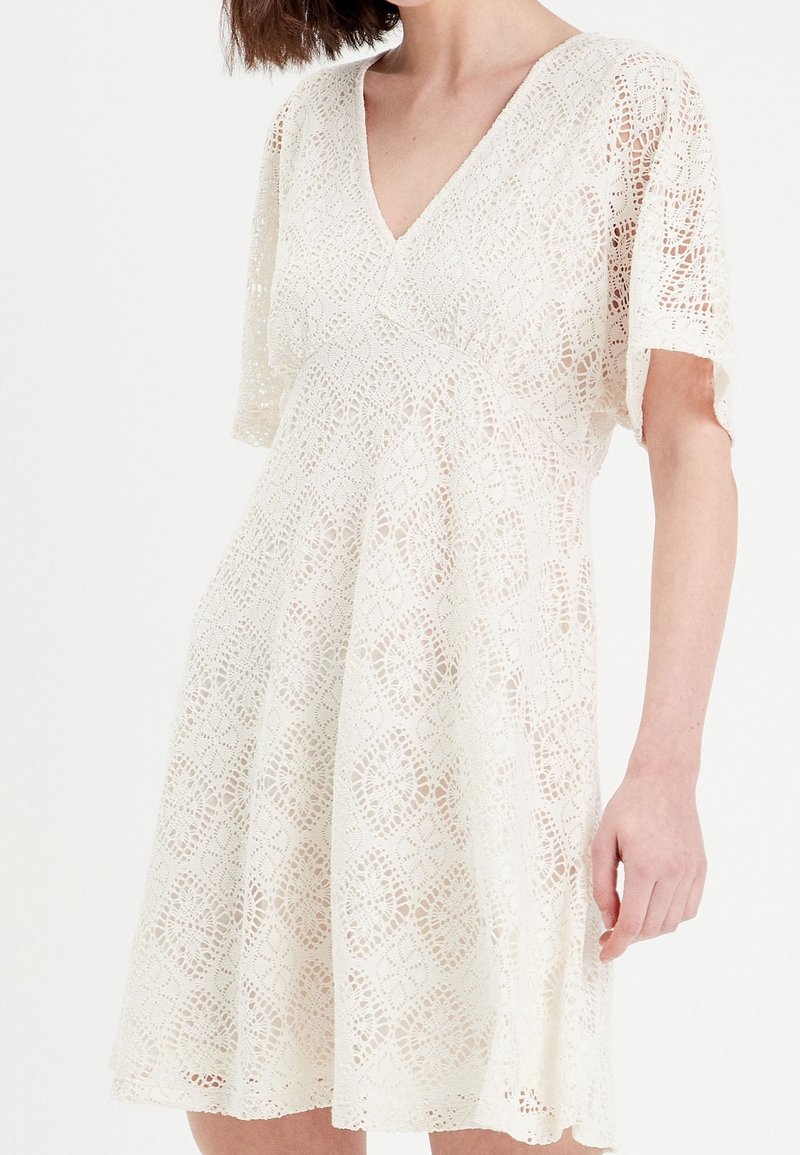 Robe blanche en dentelle à manches courtes avec un décolleté en V, ornée d'un motif floral, coupe ample, longueur genou avec une apparence texturée.