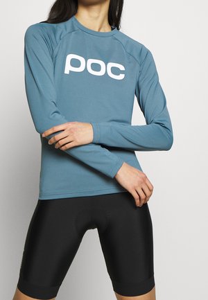 Osoba w niebieskiej sportowej koszulce z długim rękawem z białym logo "poc" oraz czarnych spodenkach kolarskich, ze skrzyżowanymi rękami na wysokości talii na jednolitym tle.