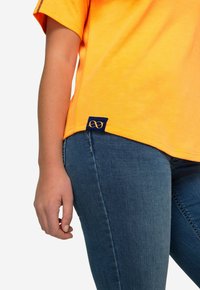 Heldoranje T-shirt met een ronde hals en korte mouwen, voorzien van een marineblauwe label aan de zoom met een gouden oneindigheidssymbool. Gedragen met een spijkerbroek.