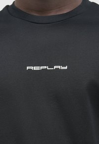 Svart T-skjorte laget av bomull. Rund halsdesign med korte ermer. Hvit "REPLAY" logo trykt på forsiden. Jevn tekstur.