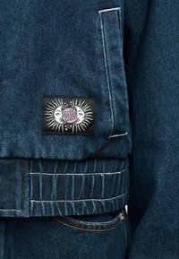 Τελείωμα τζιν μπουφάν με ελαστική ταινία και μαύρο έμπλαστρο που απεικονίζει ήλιο και φεγγάρι με το κείμενο «Santa Cruz» σε ροζ και λευκό.