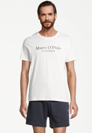 Mand med skæg iført en hvid Marc O’Polo t-shirt og sorte shorts, stående foran en ensfarvet baggrund.