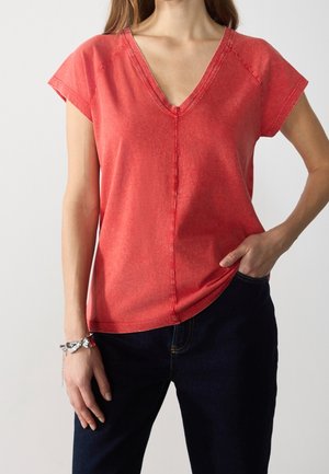 Vrouw die een vervaagde rode V-hals T-shirt met korte mouwen draagt en een donkere broek, één hand in de zak en een zilveren armband om de andere pols.