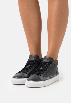 Benen met zwarte hoge sneakers met witte zolen en zwarte veters, met het "G-STAR RAW" logo aan de zijkant.