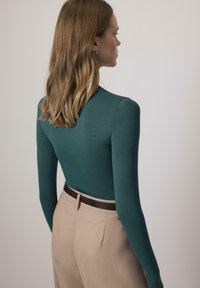 Naine, kellel on õlgadeni ulatuvad helepruunid juuksed, kannab türkiissinist pikka varrukatoppi, mis on sisse pandud beežidesse pükstesse tume vööd kandes, seisab külg ees.