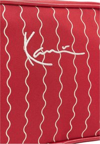 Červená textilní taška se zvlněnými bílými stehmi a vyšívaným podpisem. Hladký povrch s pevným designem.
