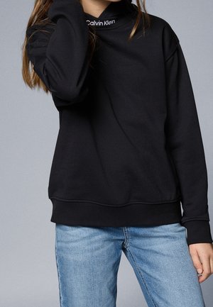 Persoon die een zwarte Calvin Klein-trui en lichtblauwe spijkerbroek draagt, met één hand het haar aanrakend tegen een effen grijze achtergrond.