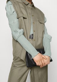 Beżowa bezrękawnik nałożona na jasnoszary, strukturalny top z długim rękawem. Trzyma czarną kopertówkę o gładkim wykończeniu.