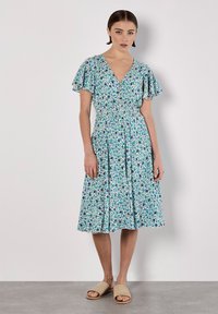 Blumenmuster-Kleid mit V-Ausschnitt, kurzen Rüschensäumen und tailliertem Bund. Die Farben umfassen Blau, Grün und Weiß mit einem fließenden Stoff.