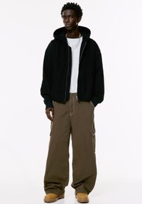 Giovane uomo che indossa un hoodie nero con zip, una maglietta bianca, pantaloni cargo marroni larghi, stivali beige e collane di catena argentate, in piedi contro uno sfondo bianco.