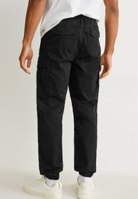 Personne portant un pantalon cargo noir avec poches à rabat et poignets élastiques, baskets blanches, debout devant un fond clair uni.