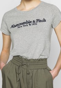 Grå kortærmet t-shirt med marineblå broderet tekst "Abercrombie & Fitch New York 1892" og en afslappet pasform, parret med grønne bukser med snor.