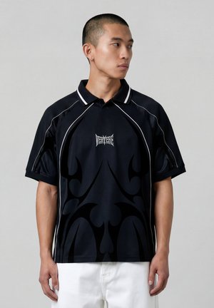 Junger Mann, der ein schwarzes Poloshirt mit weißen Paspeln und abstrakten dunklen Mustern trägt, kombiniert mit weißen Hosen, schaut zur Seite.