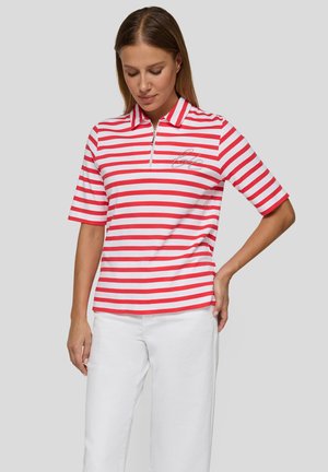 Frau trägt ein rot-weiß horizontal gestreiftes kurzärmeliges Poloshirt mit Reißverschluss und weiße Hose, steht mit einer Hand in der Hüfte.