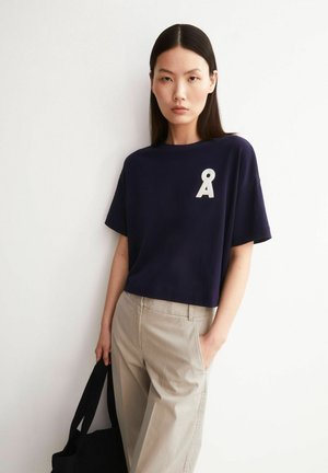 Femme aux cheveux lisses et foncés portant un t-shirt bleu marine avec un logo blanc Å, un pantalon beige, tenant un sac noir, debout contre un mur blanc.