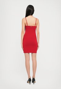 Vestito rosso a costine con spalline sottili, silhouette aderente e dettaglio di chiusura sul retro. Indossato con scarpe nere con tacco alto.