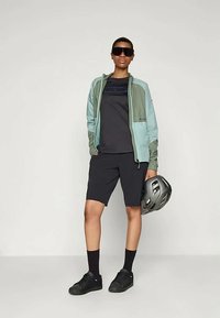 Αθλητική εμφάνιση που περιλαμβάνει ένα ελαφρύ πράσινο και γκρι φούτερ με φερμουάρ, μαύρη μπλούζα, μαύρη βερμούδα, μαύρες κάλτσες και μαύρα αθλητικά παπούτσια. Κρατάει ένα κράνος.