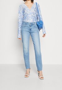 Ljusblå mönstrad transparent blus i kombination med ljusblå jeans och vita högklackade sandaler. En blå axelväska ger kontrast.