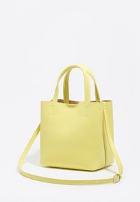 Mała, strukturalna, żółta skórzana torebka z dwoma górnymi rączkami i długim, regulowanym paskiem na ramię na białym tle.
