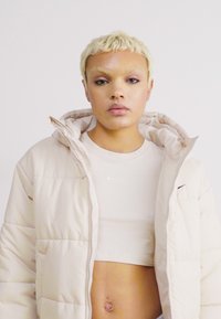 Casaco puffer de tom bege claro com fecho éclair à frente e capuz grande. Por baixo, uma t-shirt cropped ajustada de tom neutro claro, com um pequeno logotipo no peito.