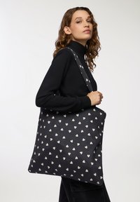 Czarna torba na ramię z białym wzorem w serca, wykonana z tkaniny, z szerokim paskiem i pojemnym, prostokątnym kształtem.