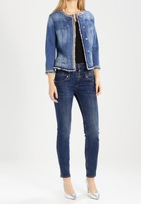 Giacca di jeans con tasche frontali e bordi decorati, abbinata a jeans scuri aderenti e tacchi alti argento, su uno sfondo neutro.