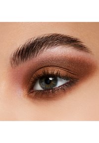 Close-up van een oog met bruin en bronzen oogschaduw, dikke donkere wimpers en een natuurlijke wenkbrauw. De iris is blauw met bruine tinten.