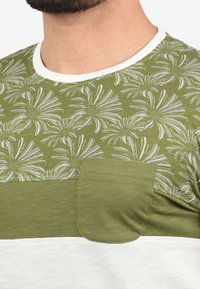 Grøn t-shirt med blomstermotiv på toppen og en ensfarvet grøn panel nedenunder. Har en kontrast hvid kant og en brystlomme.