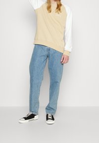 Sudadera de manga larga beige y blanca con puños acanalados, combinada con jeans de mezclilla azul claro y zapatillas negras y blancas.