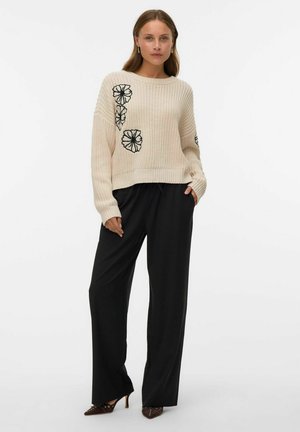 Maglione lavorato a maglia beige con ricami floreali neri, abbinato a pantaloni neri larghi e scarpe con tacco alte marroni. Scollo rotondo semplice.