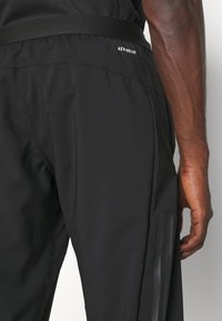 Svarta sportbyxor har ett mjukt tyg, elastisk midja och tre vertikala ränder på vänster ben. Texten lyder "AEROREADY."