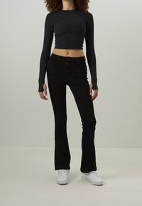 Femme portant un crop top noir à manches longues et un pantalon évasé noir avec des baskets blanches, debout devant un fond clair uni.