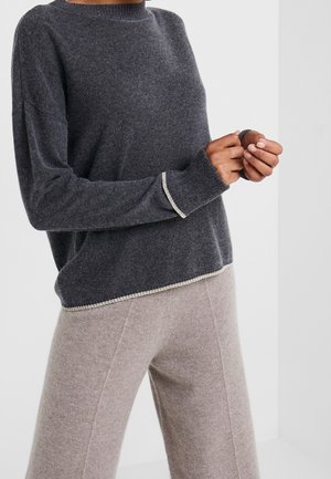 Osoba nosící tmavě šedý svetr s dlouhým rukávem s bílým lemováním a světle béžové kalhoty se širokými nohavicemi na jednobarevném pozadí.