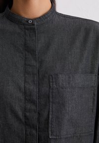 Camicetta grigia scura con bottoni, realizzata in tessuto testurizzato, dotata di colletto alla cinese e di una tasca sul petto.