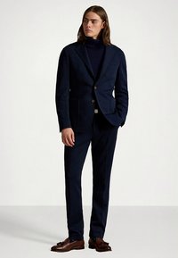 Granatowy garnitur z dopasowaną marynarką i pasującymi spodniami, posiadający klapę z wycięciem, dwie przednie kieszenie oraz brązowe skórzane buty.