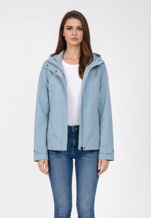 Kobieta w jasnoniebieskiej kurtce z kapturem na białej koszuli i niebieskich dżinsach, stojąca na jednolitym białym tle.