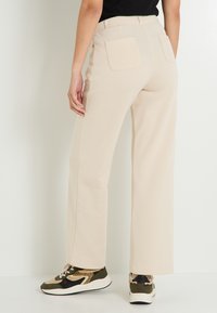 Pantalones de pierna ancha beige con cintura alta, que cuentan con dos bolsillos traseros. Combinados con zapatillas estampadas en tonos oliva, negro y beige.