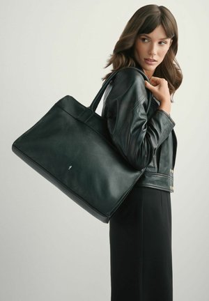 Kobieta w czarnej skórzanej kurtce trzyma dużą czarną skórzaną torbę na ramię na jednolitym tle.