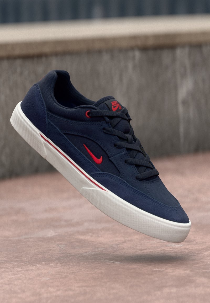Baskets Nike bleu foncé avec logo rouge et semelle blanche, montrées en l'air au-dessus d'une surface pavée et d'un fond en béton.