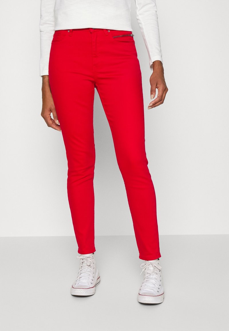 Femme portant un pantalon slim rouge vif, un haut à manches longues blanc et des baskets montantes blanches, se tenant devant un fond uni.