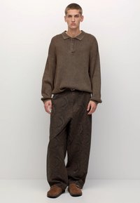 Brun stickad polo-shirt med krage och tre knappar, ihop med löst sittande mörka mönstrade byxor. Bruna slipskor fulländar outfiten.