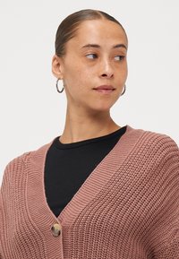 Cardigan de punto color rosa con escote en V, que presenta un cierre de un solo botón y textura acanalada, usado sobre una blusa negra.
