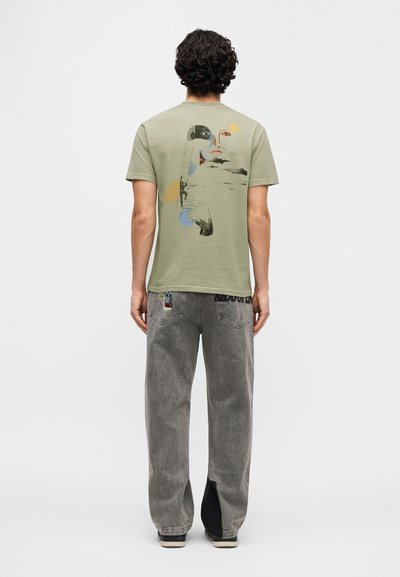 Λαδί πράσινο t-shirt με αφηρημένα γραφικά στην πλάτη, συνδυασμένο με γκρι τζιν denim που διαθέτει πάνελ αντίθεσης και λεπτομέρειες με μπαλώματα.