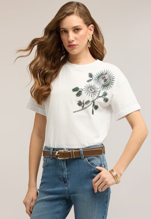Mujer con cabello largo y ondulado lleva una camiseta blanca con bordado floral verde, jeans azules, cinturón marrón, pendientes dorados y pulseras sobre un fondo liso.