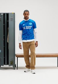 Blaues Sporttrikot mit geometrischen Mustern, darunter ein weißes langärmliges Hemd, hellbraune Hose und weiße Sneakers. Hintergrund mit einem Spind.